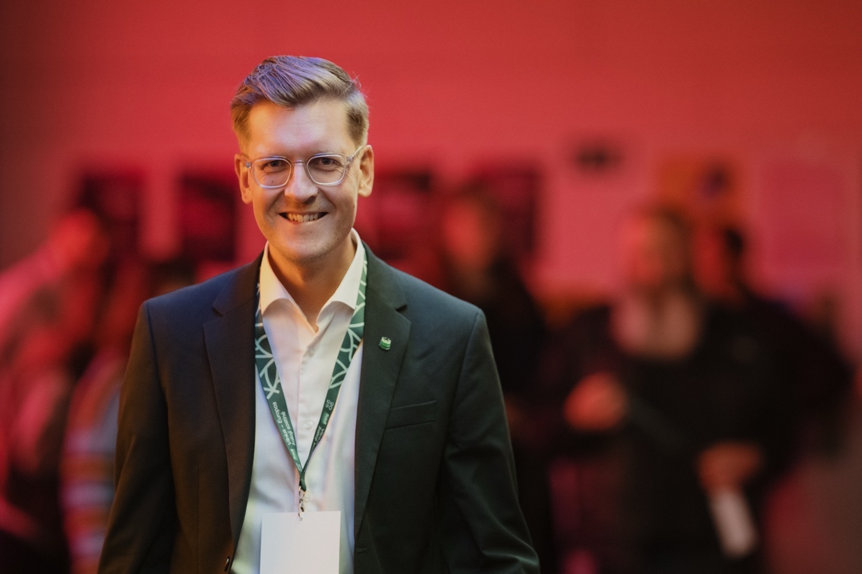 A man in a suit smiles at the camera. He is wearing glasses and a lanyard.