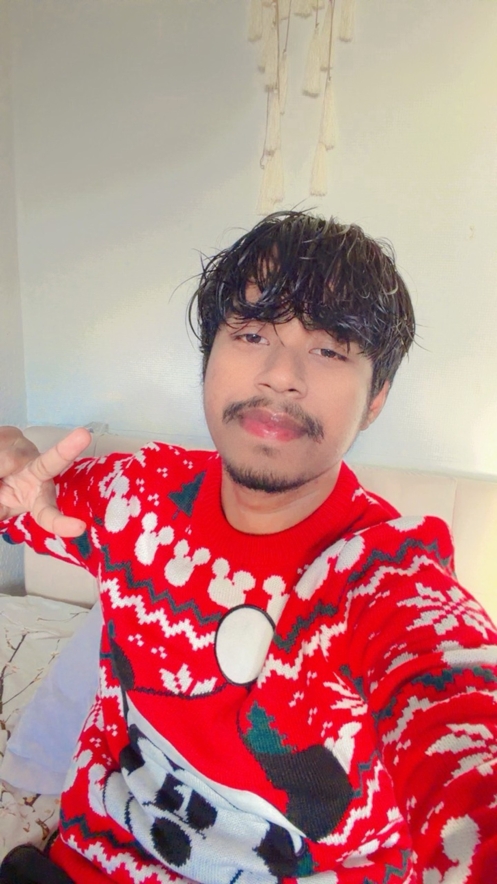 A man poses for a selfie, wearing a festive red Christmas sweater. He flashes a peace sign at the camera.