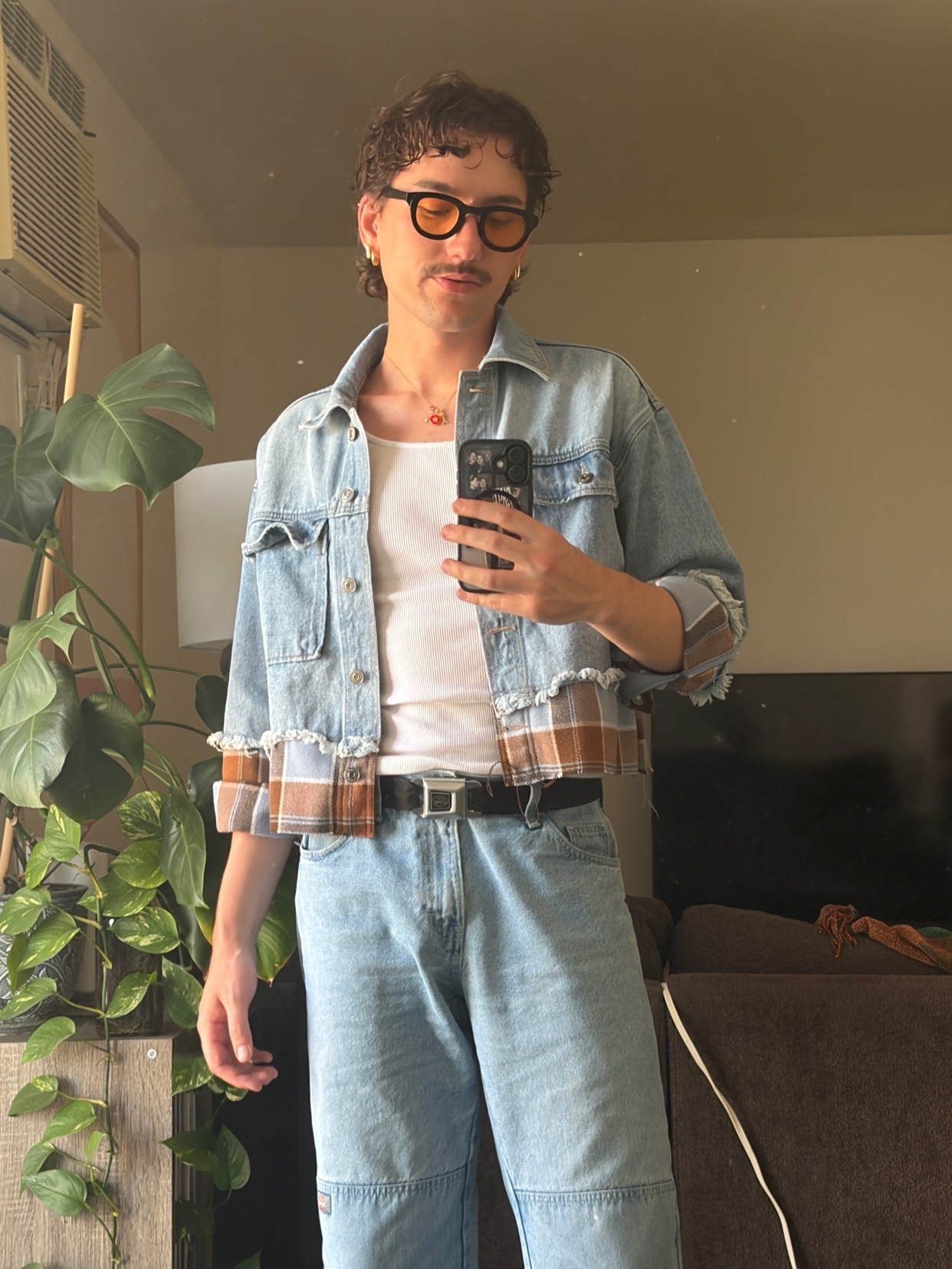A man with a mustache and curly hair takes a selfie in a denim jacket and sunglasses. He is wearing a white tank top and light wash jeans.