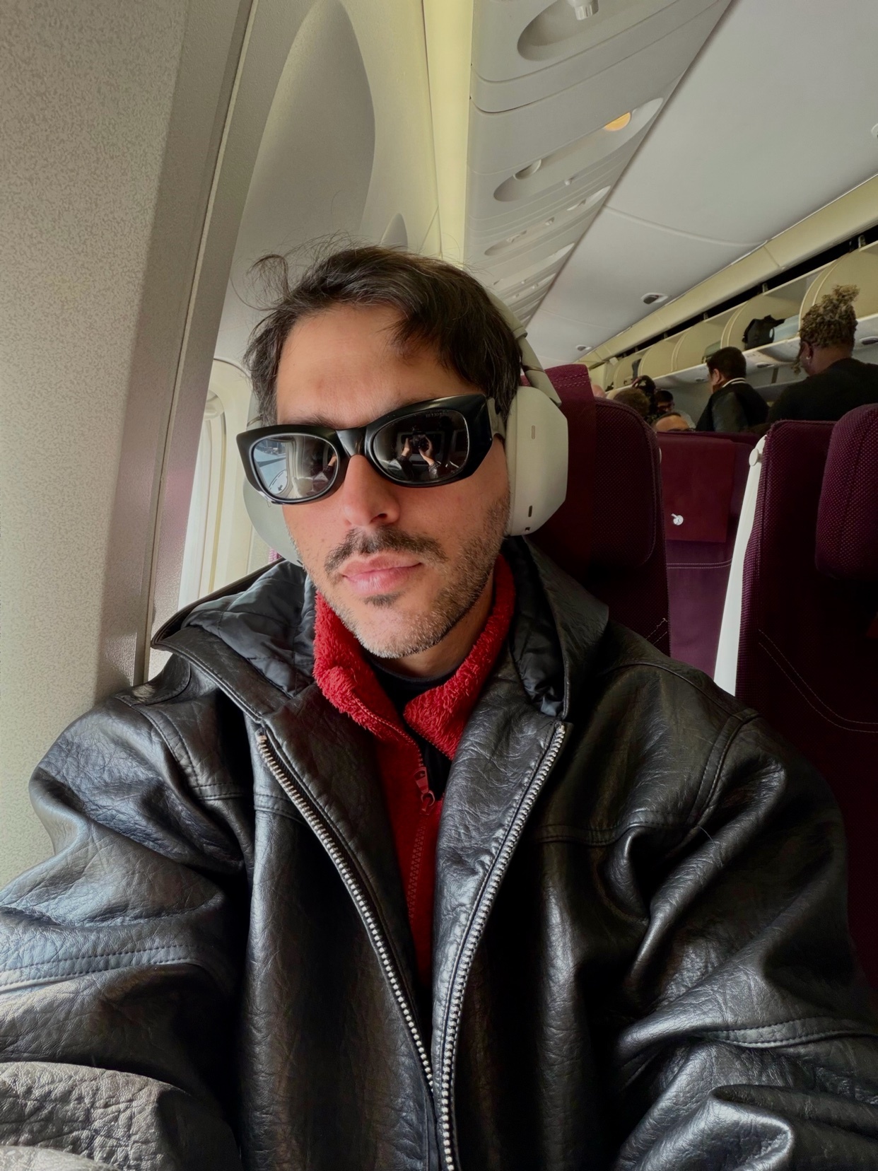 A man wearing sunglasses and headphones on an airplane. He is wearing a leather jacket and looking at the camera.