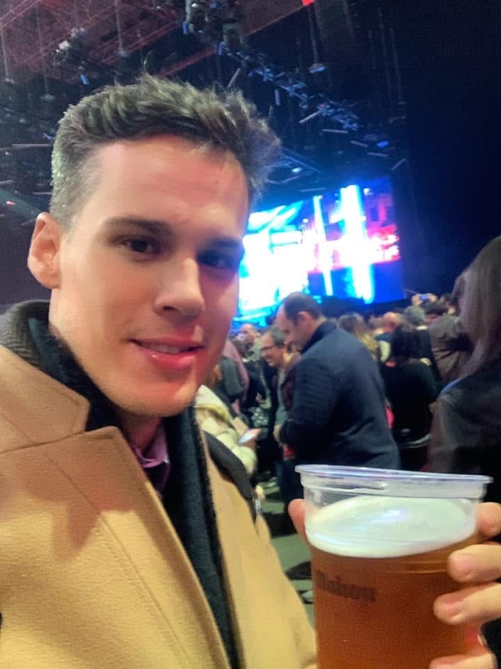 A man takes a selfie while holding a beer at an event with a large crowd in the background.