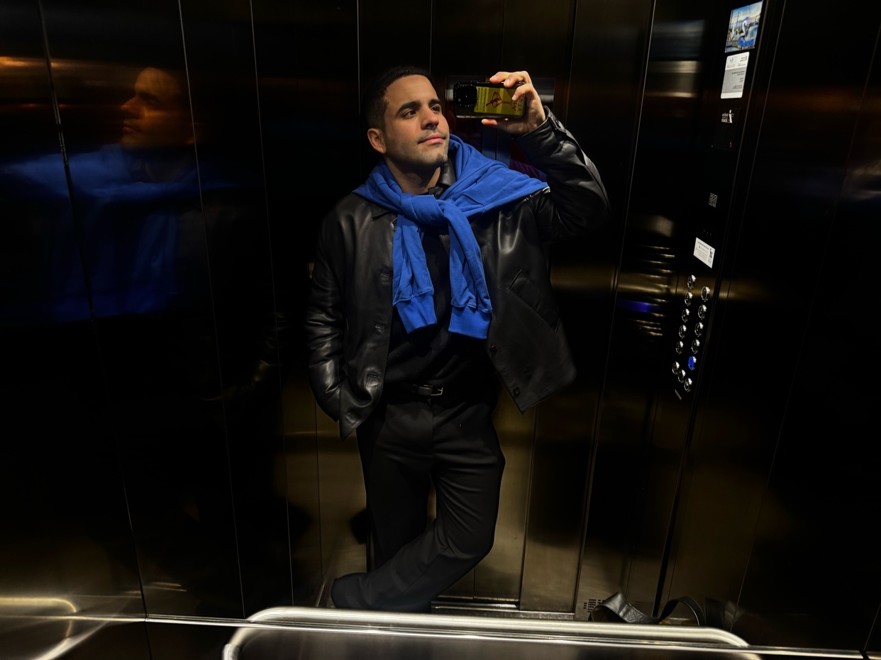 A man takes a selfie in an elevator. He is wearing a black leather jacket and a blue scarf.
