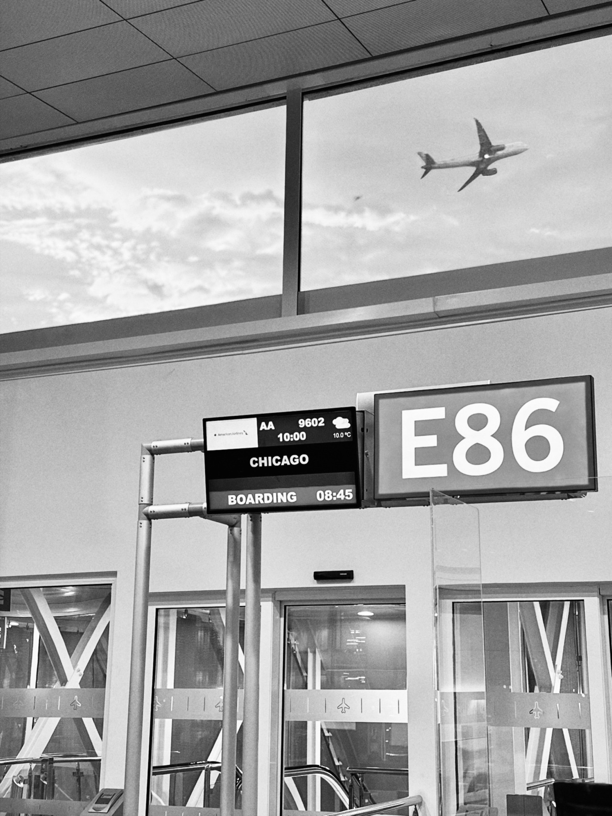 A black and white photo of an airport gate with a departure board showing a flight to Chicago. An airplane is visible in the sky outside the window.
