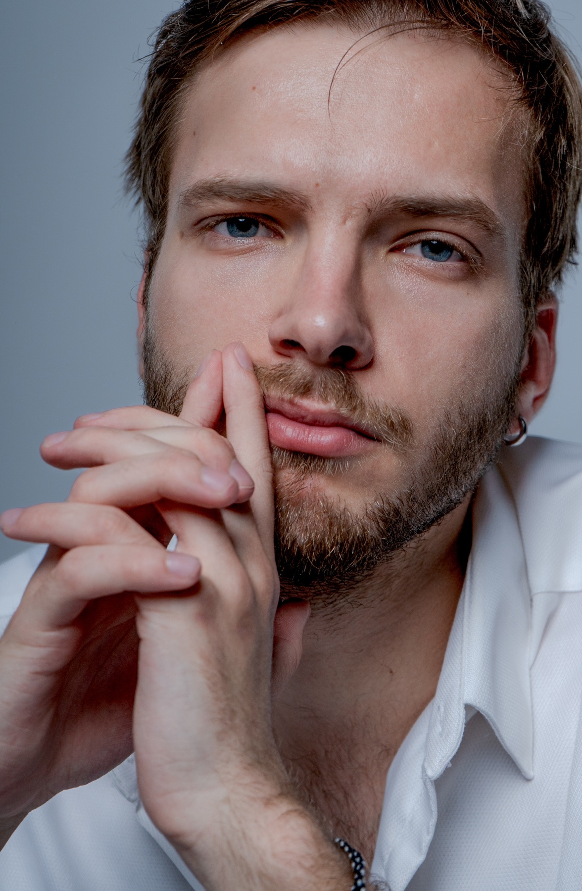A close-up portrait of a man with stubble and a beard, looking directly at the camera with his hands clasped near his face.
