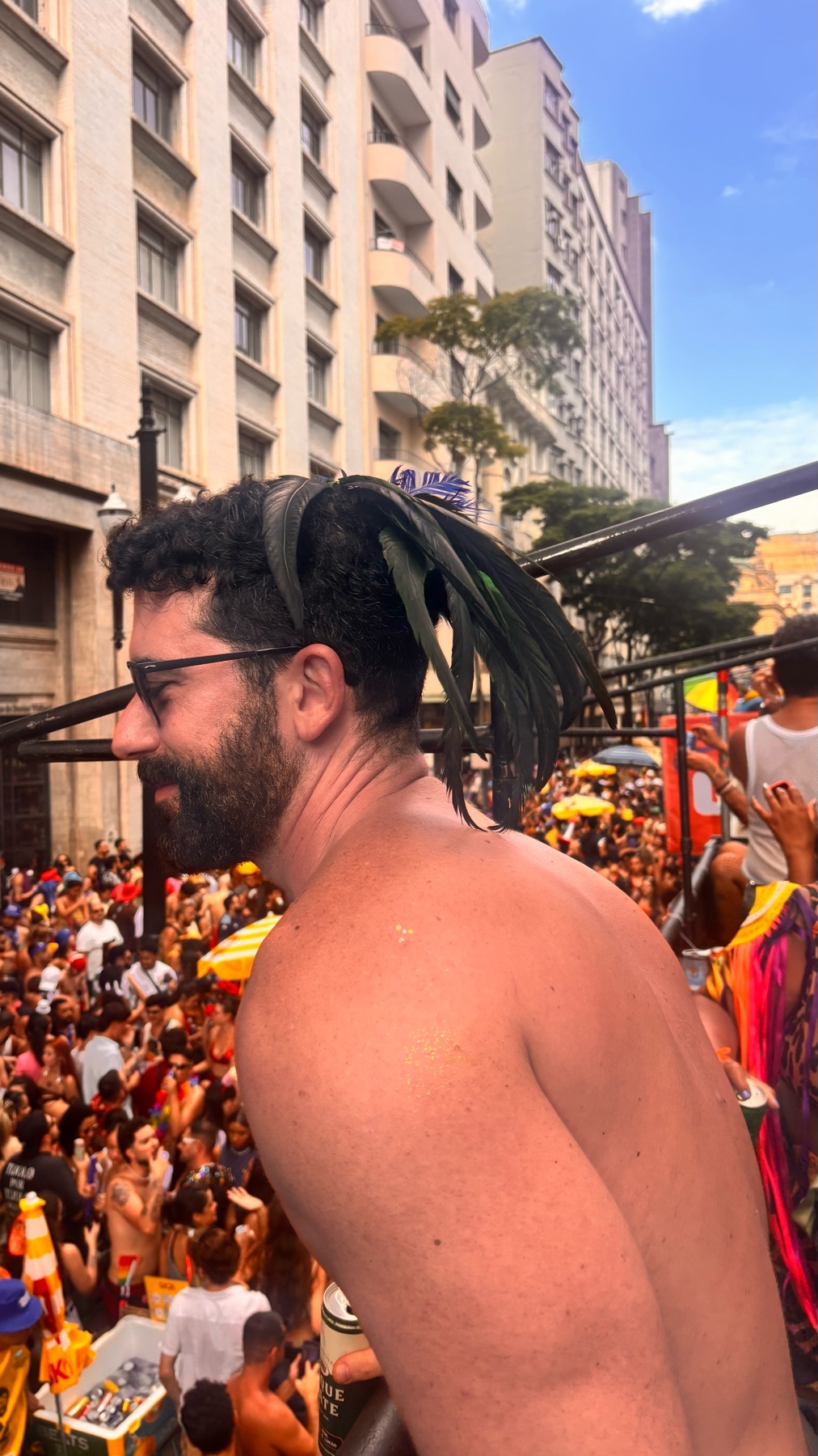 A man with a beard and sunglasses is seen from the side, wearing feathers in his hair. He is shirtless and appears to be in a crowded street celebration, possibly a carnival or parade.