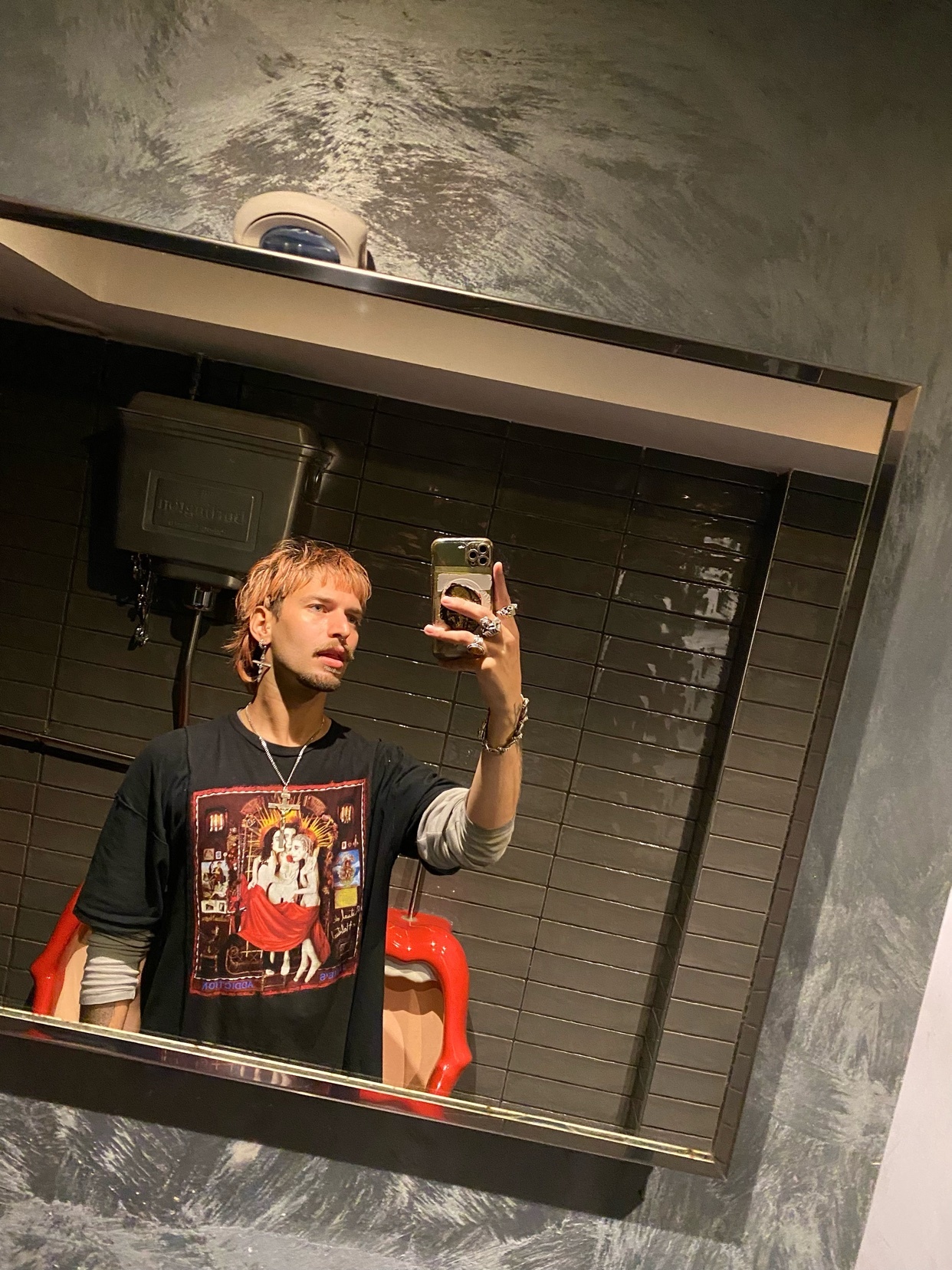 A man takes a mirror selfie in a bathroom. He is wearing a graphic t-shirt and has blonde, curly hair.