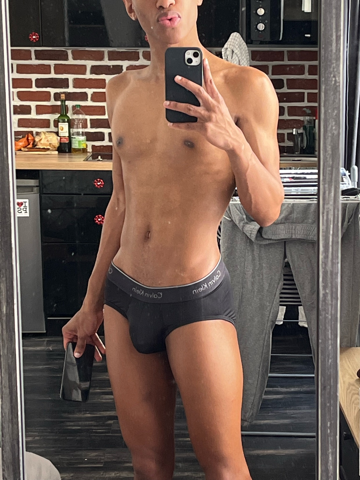 A man in black Calvin Klein briefs takes a mirror selfie in a kitchen. He is holding a black bottle in his left hand and posing with his lips puckered.
