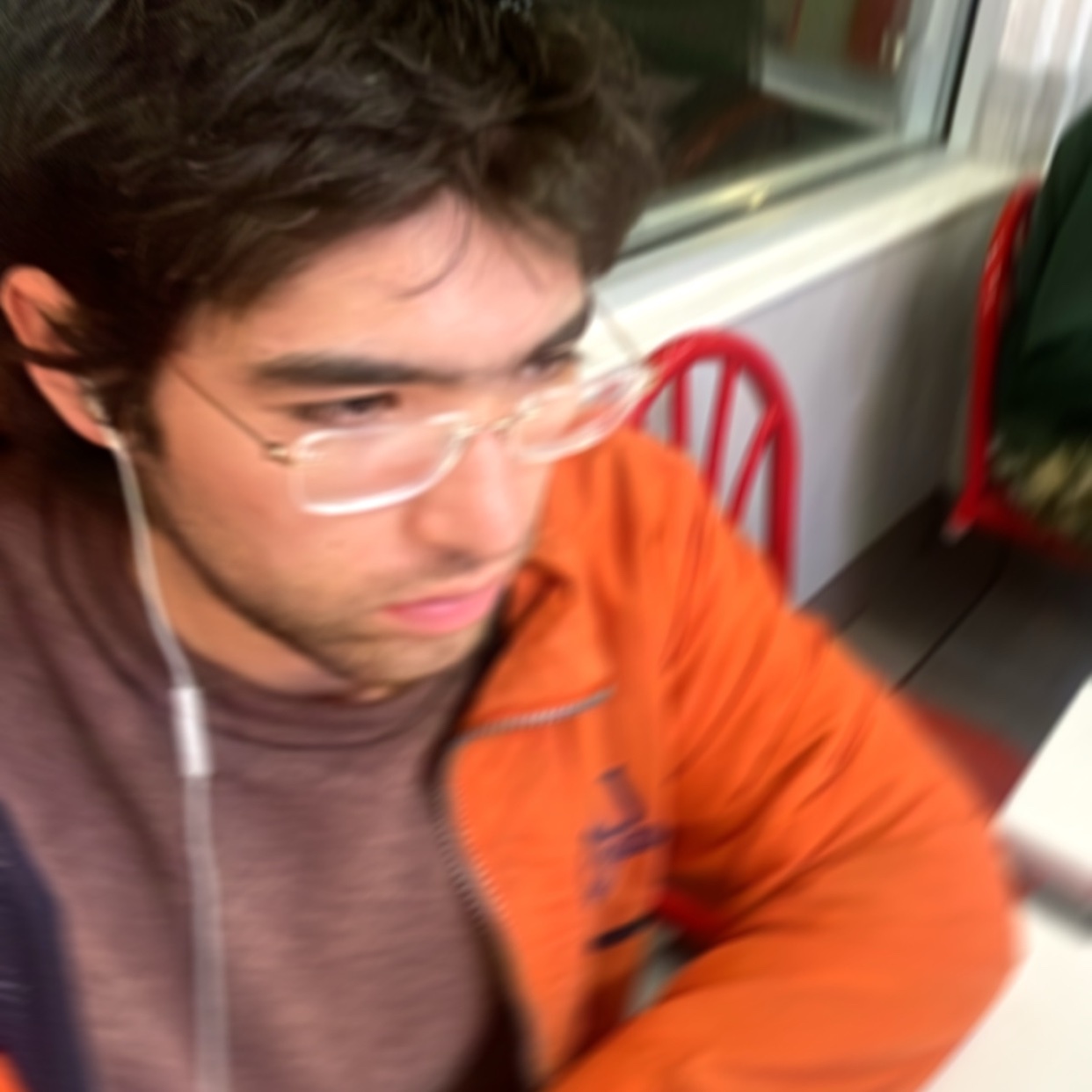 A blurry photo of a man wearing glasses and an orange jacket. He appears to be indoors, possibly at a restaurant or cafe.