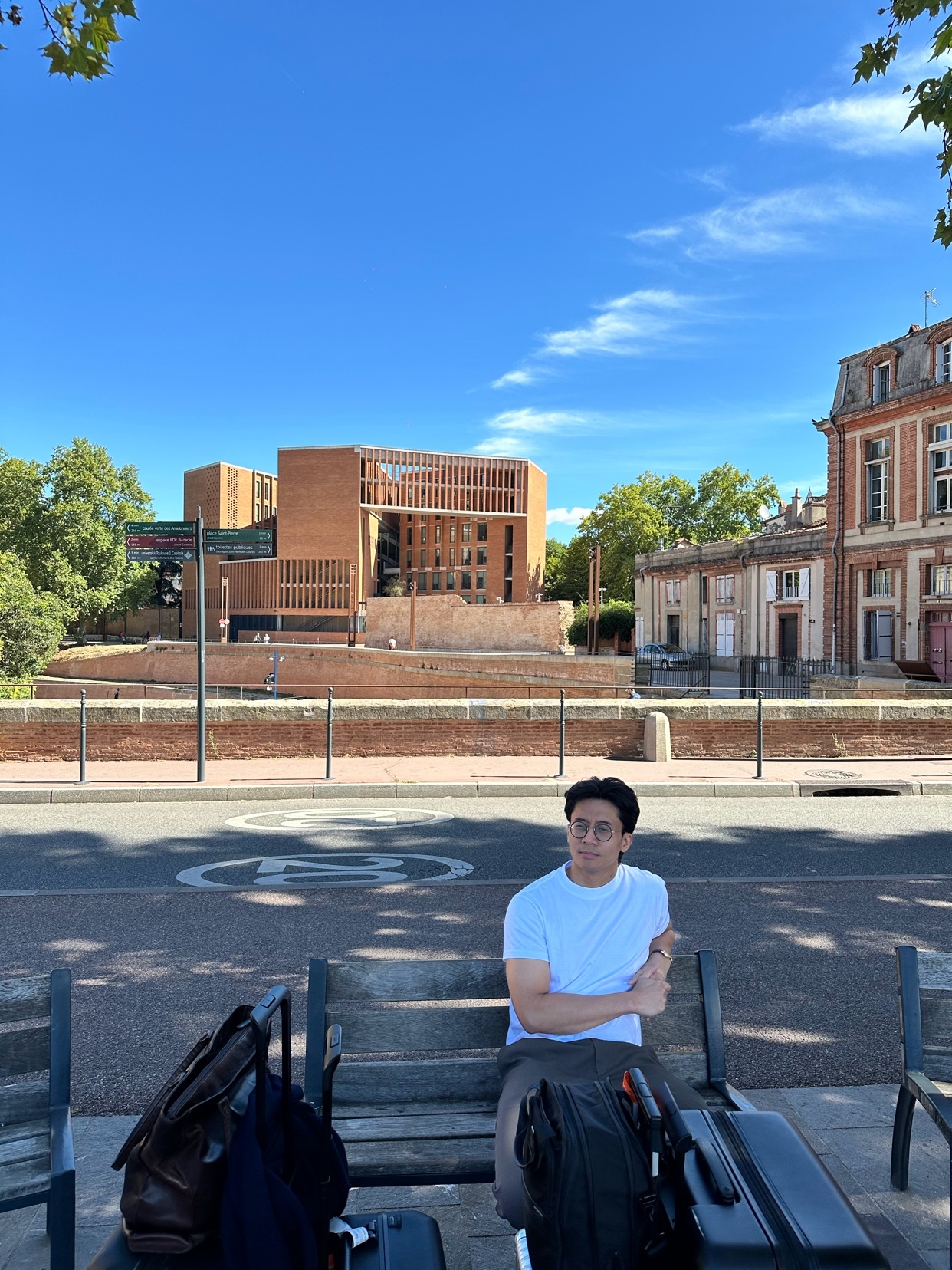 A man sits on a bench with luggage beside him, looking towards the camera. Behind him, modern brick architecture stands against a clear blue sky.