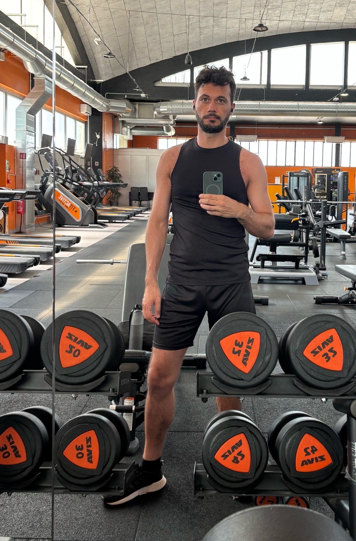 A man in a black tank top and shorts takes a mirror selfie in a gym, surrounded by dumbbells and exercise equipment.