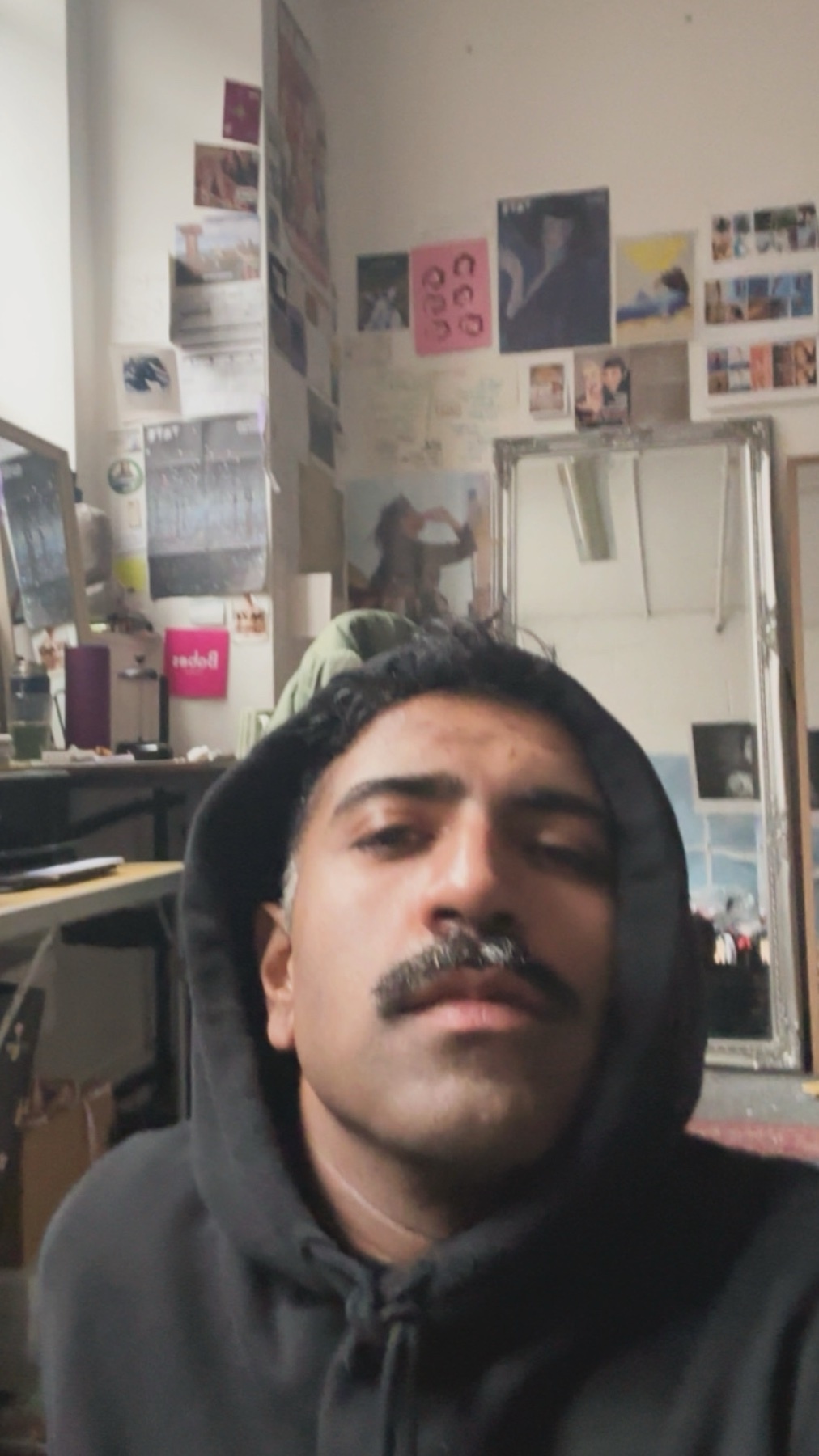 A man takes a selfie in his bedroom. The room is decorated with posters and a mirror.