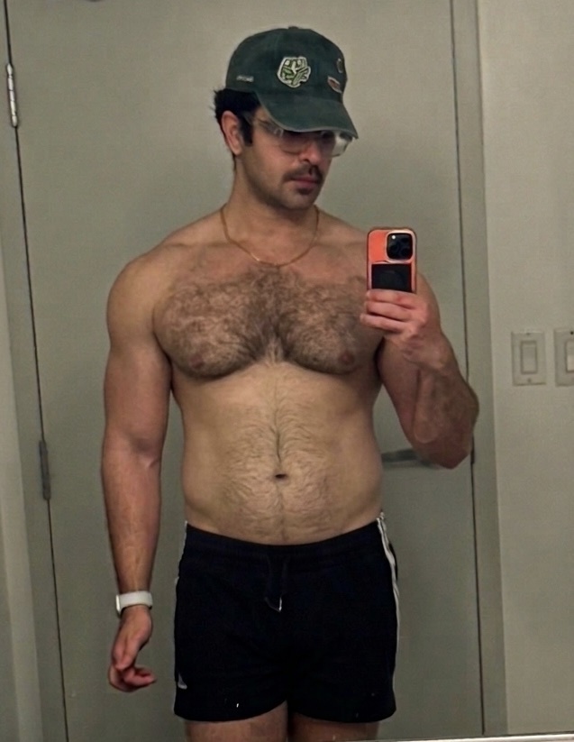 A man with a muscular physique and significant chest hair poses for a mirror selfie. He is wearing a baseball cap and shorts, with a phone in his hand.