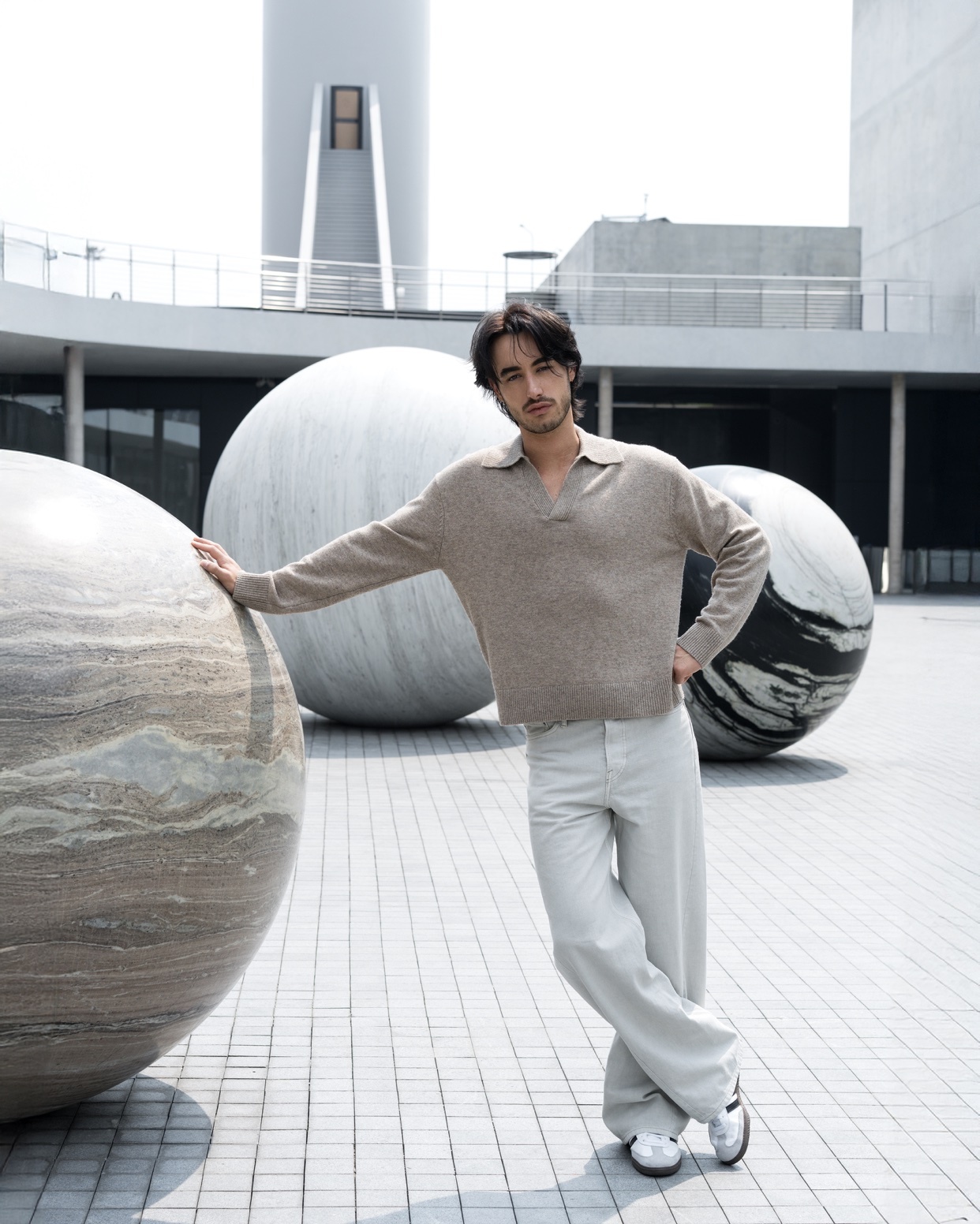 A man poses confidently in front of large, abstract spherical sculptures and modern architecture. He is wearing a light brown collared sweater and light grey wide-leg pants, exuding a stylish and casual demeanor.