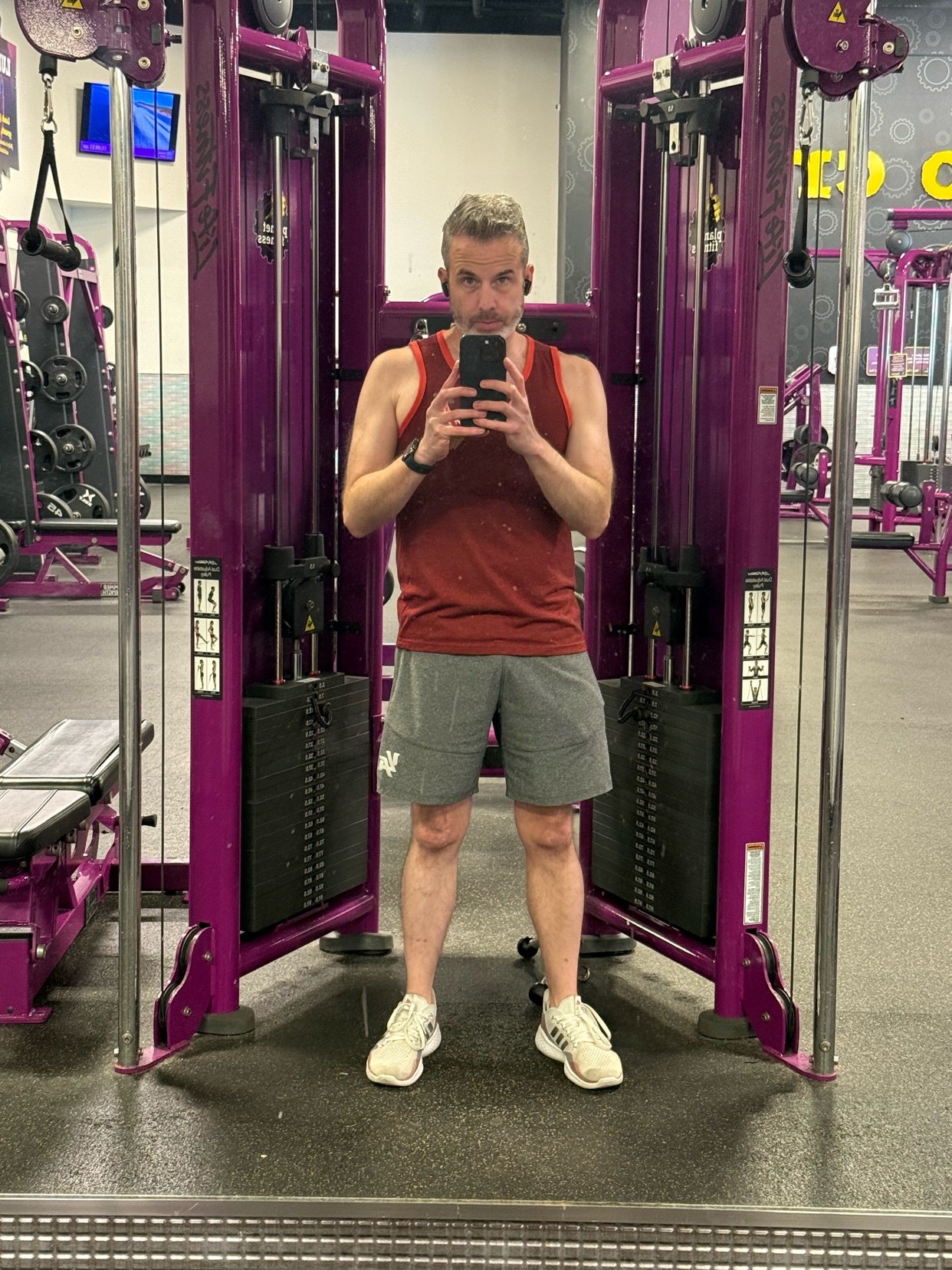 A man takes a mirror selfie at the gym. He is wearing a tank top and shorts.