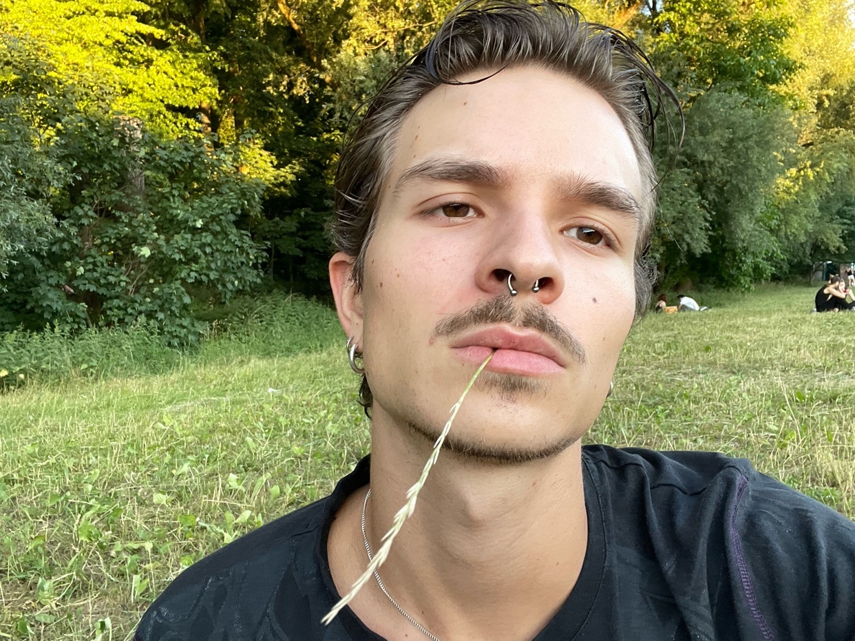A man poses for a selfie outdoors, with a piece of grass in his mouth. The photo is taken in a grassy area with trees in the background.