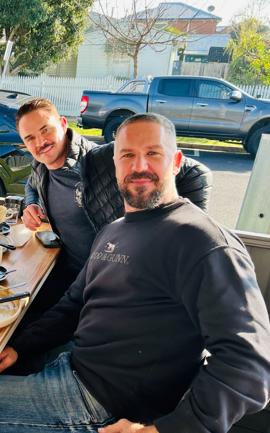 Two men pose for a photo outdoors during the day. The man in the foreground wears a black sweatshirt and jeans, while the man behind him wears a puffer vest over a dark t-shirt and jeans.