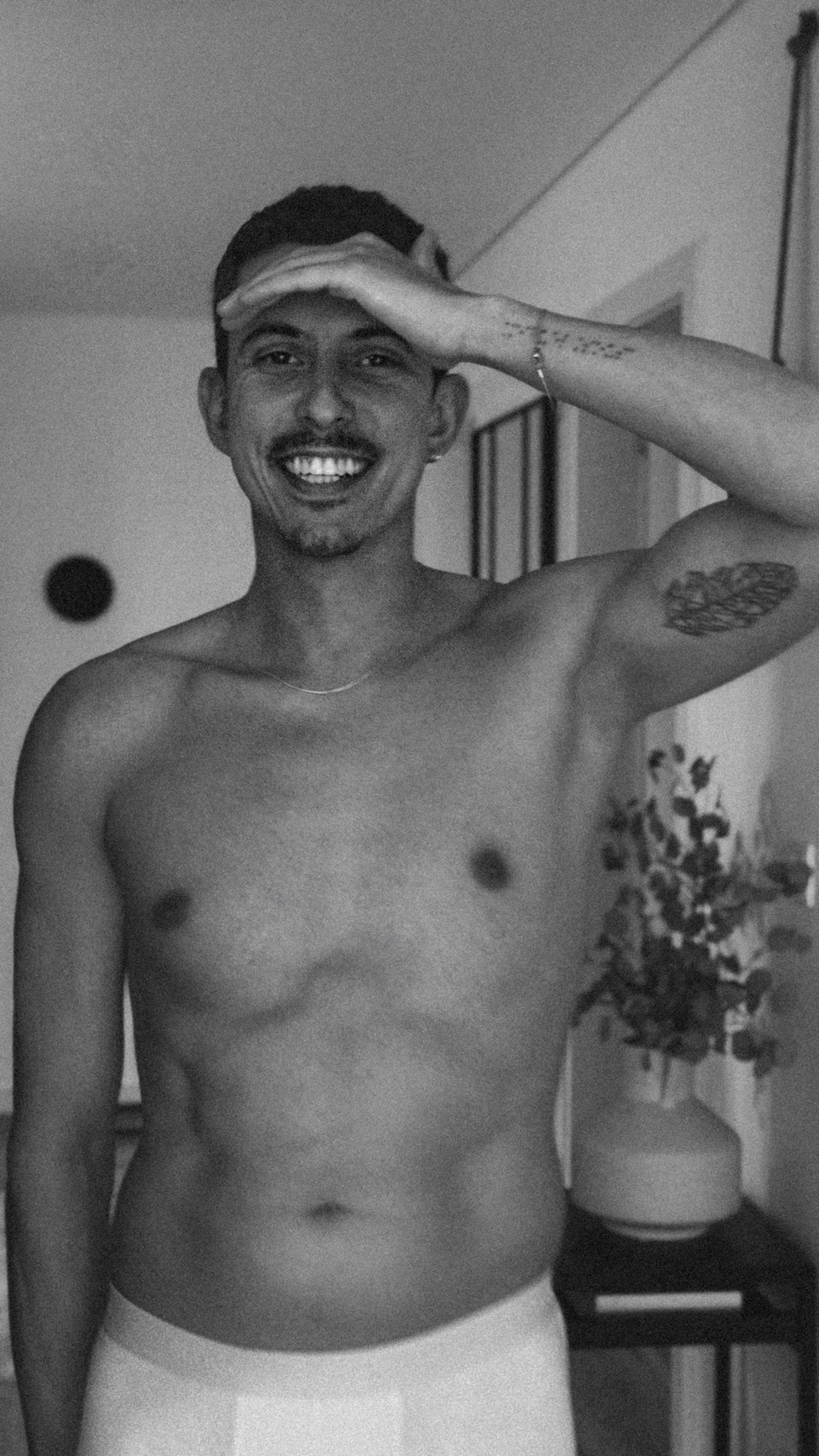 A black and white photo of a man wearing underwear and no shirt, smiling and looking towards the camera with his hand shielding his eyes.