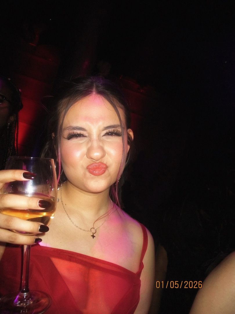 A young woman with dark hair and dramatic makeup poses for a photo while holding a glass of wine. She is wearing a red dress and making a funny face.