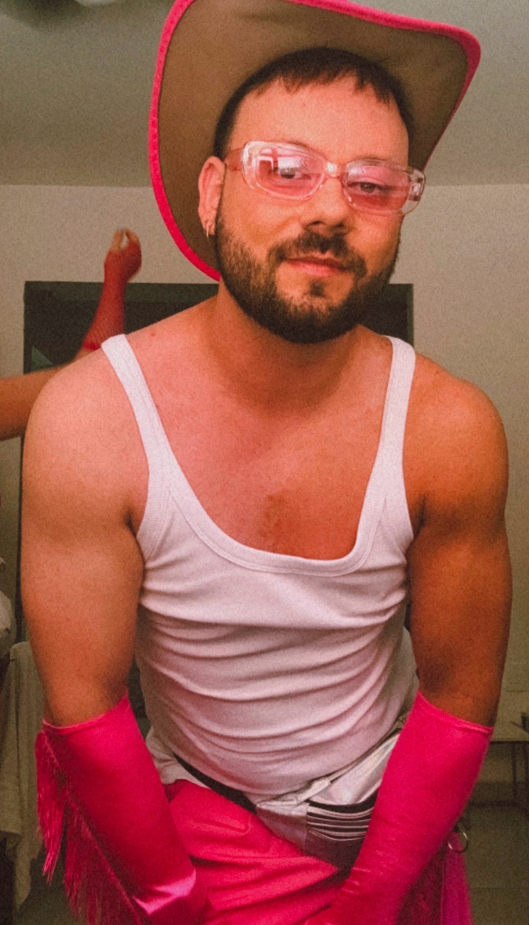 A man in a cowboy hat, pink glasses, and gloves poses for a selfie. He is shirtless, adding a touch of sensuality to the carnival-themed photo.