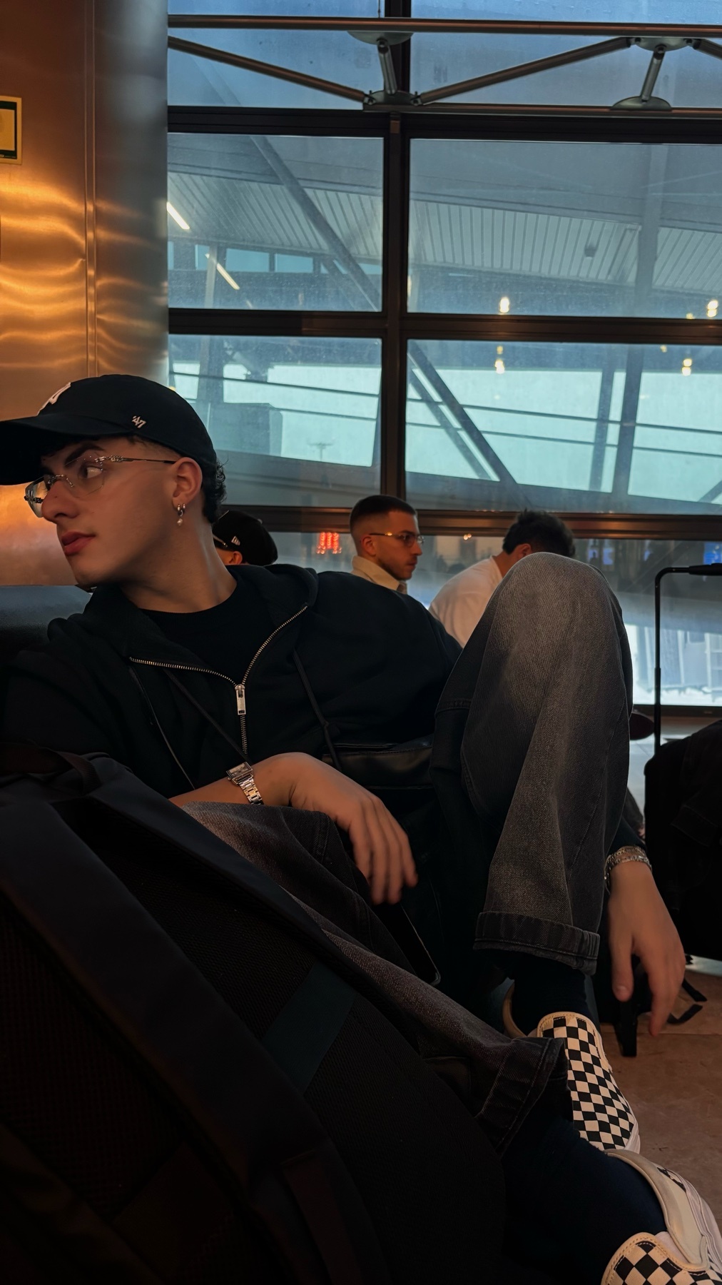 A young man wearing a baseball cap and glasses sits in an airport lounge, looking off to the side. He is dressed casually in a zip-up hoodie and jeans, with a backpack beside him.