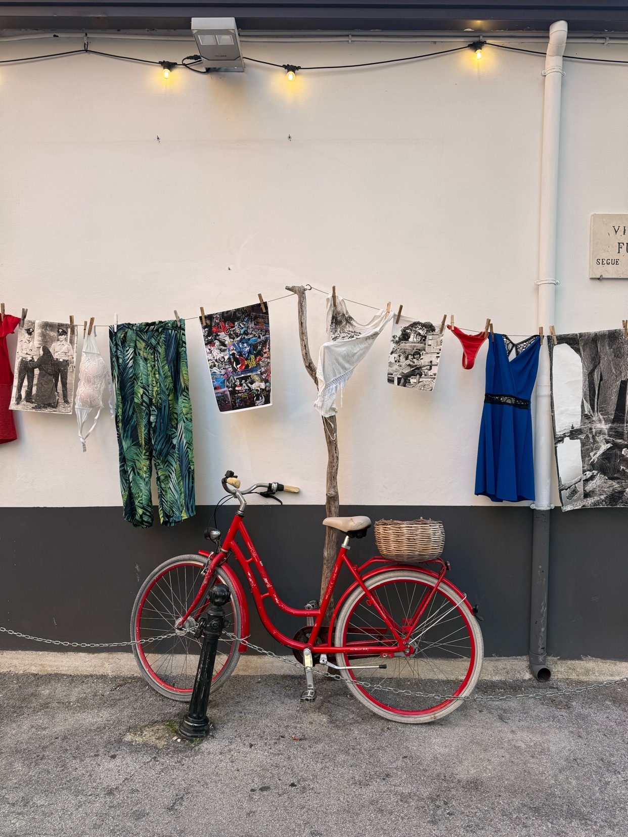 A vibrant red bicycle with a wicker basket is parked in front of a white wall adorned with a clothesline displaying various garments. String lights overhead add a whimsical touch to this charming street scene.