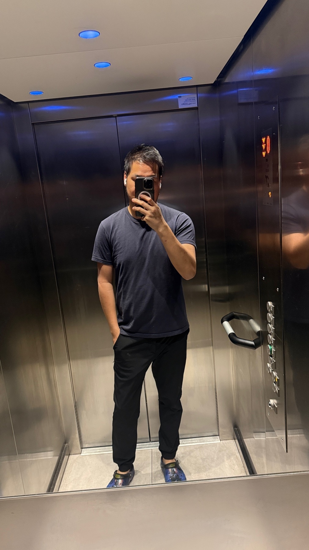 A man takes a selfie in an elevator. He is wearing a dark t-shirt and black pants.