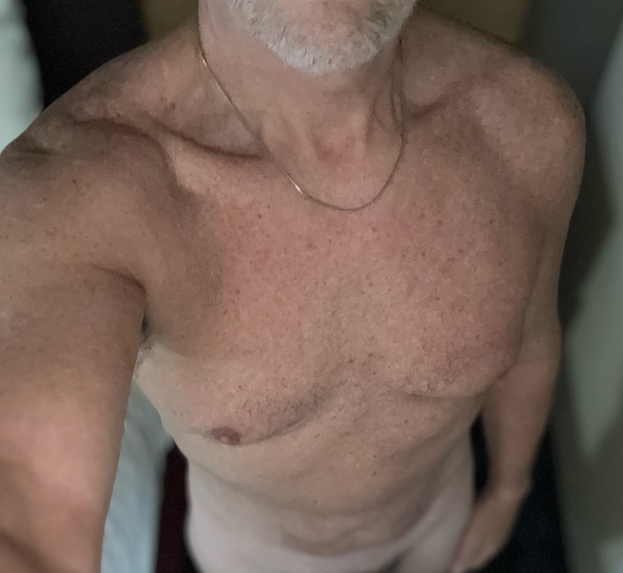 A man with grey stubble and grey hair is shown from the chest up, wearing a thin gold chain. He is shirtless and appears to be in a private setting.