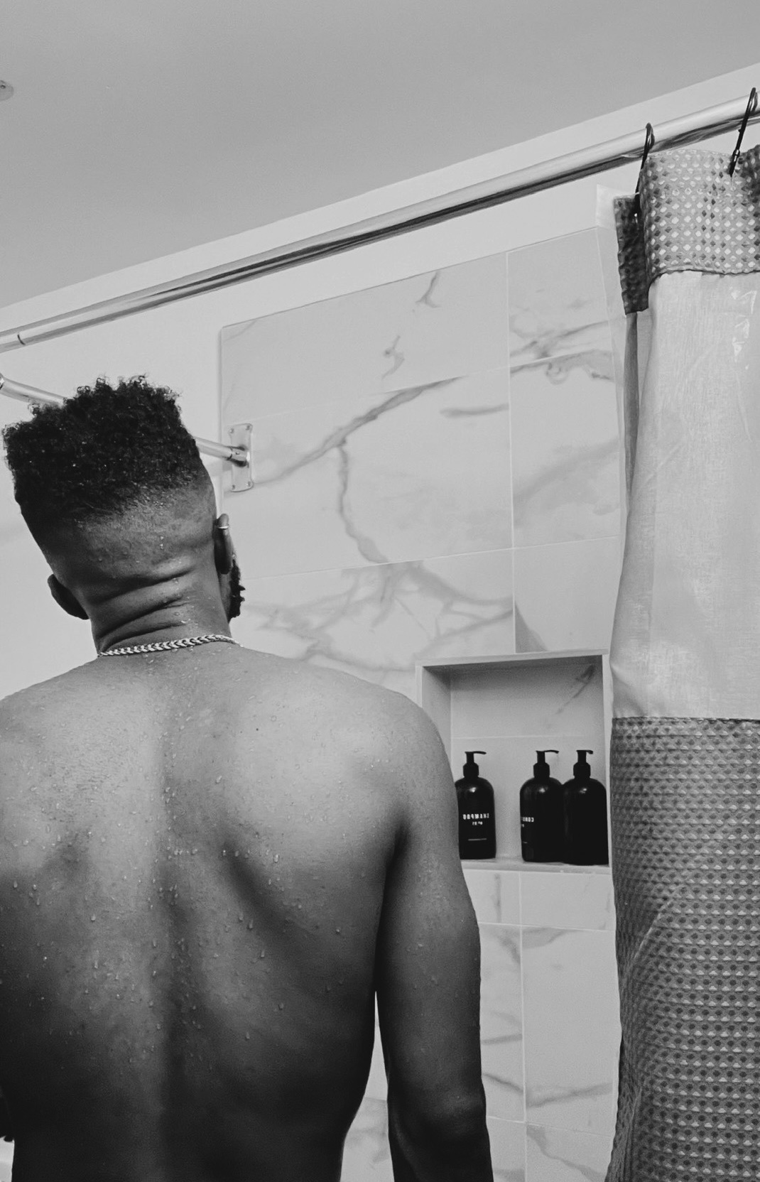 A man stands shirtless in a modern shower. The black and white photo highlights his muscular back and the sleek bathroom design.