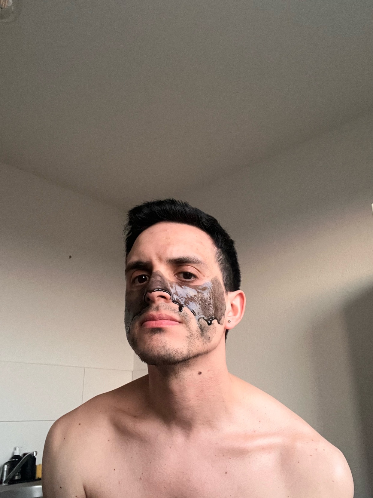 A man with dark hair and a peel-off face mask applied to his nose and cheeks looks directly at the camera. He is shirtless in what appears to be a bathroom.