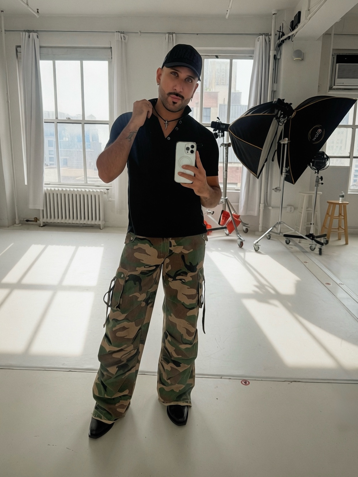 A man in a black polo shirt, camouflage cargo pants, and a black baseball cap takes a selfie in a photography studio. He is adjusting the collar of his shirt and holding a smartphone.