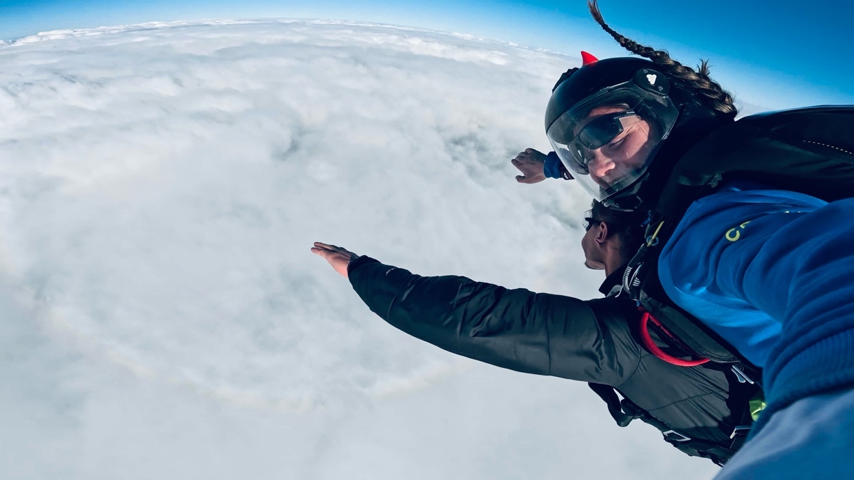 Two people are skydiving together, with one person in front and the other behind them. They are both wearing helmets and goggles, and are surrounded by clouds.