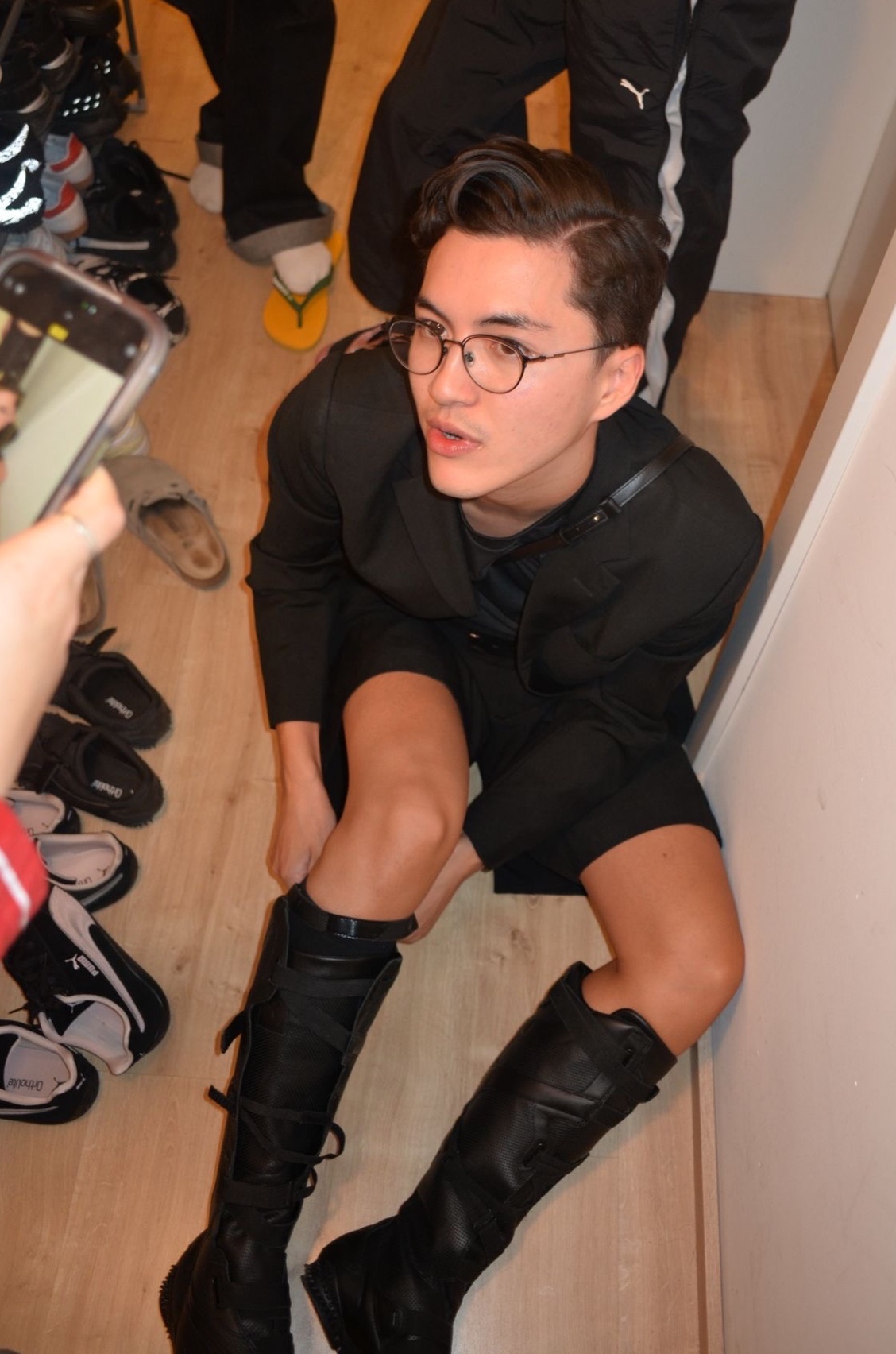 A man wearing glasses and black boots poses for a photo. He is surrounded by other people and shoes.