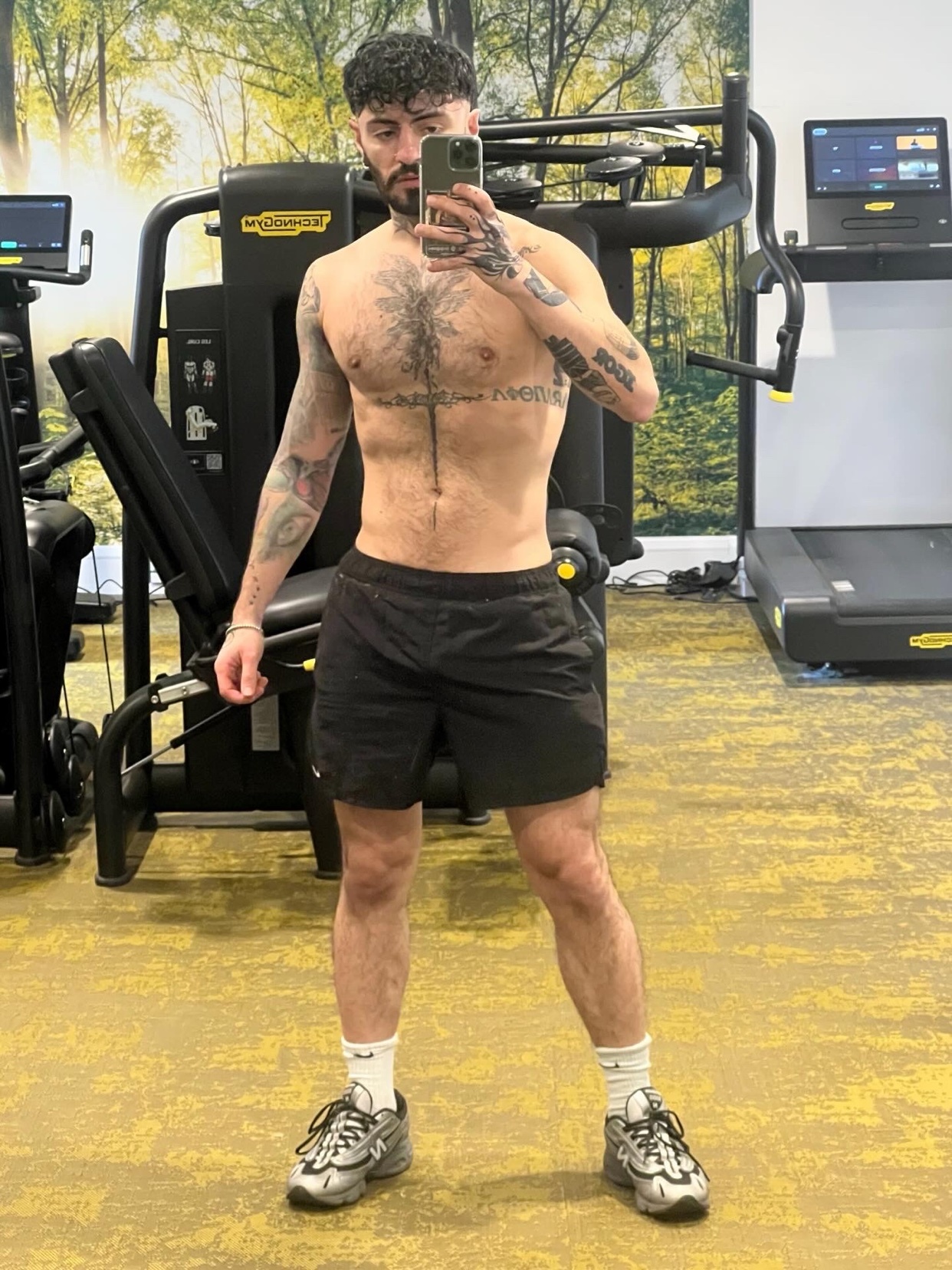 A man with tattoos on his arms and chest takes a selfie in a gym. He is wearing black shorts and athletic shoes.