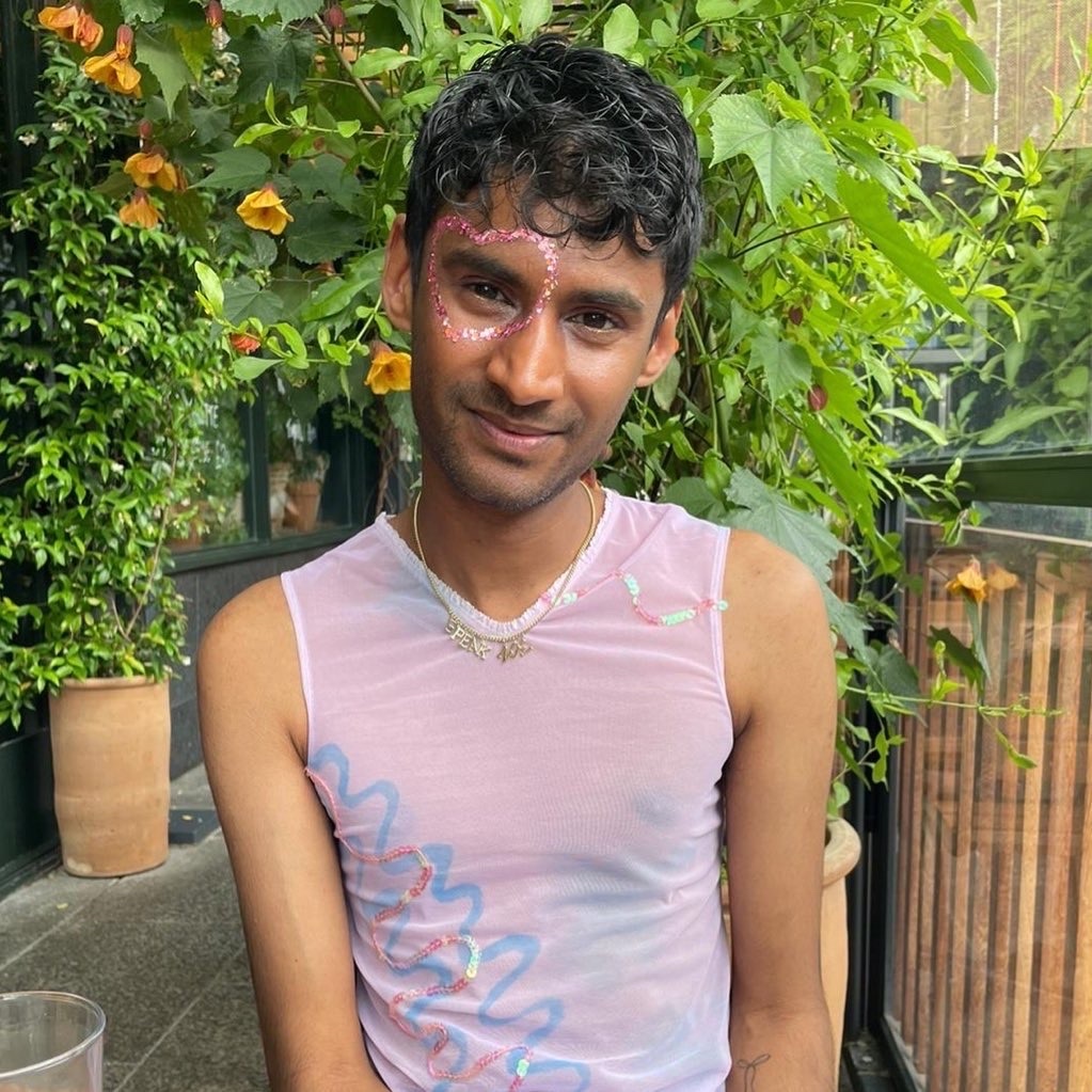 A person with glitter makeup around their eyes smiles at the camera, wearing a sheer pink tank top with blue and pink wavy lines. They are seated outdoors with lush green plants in the background.