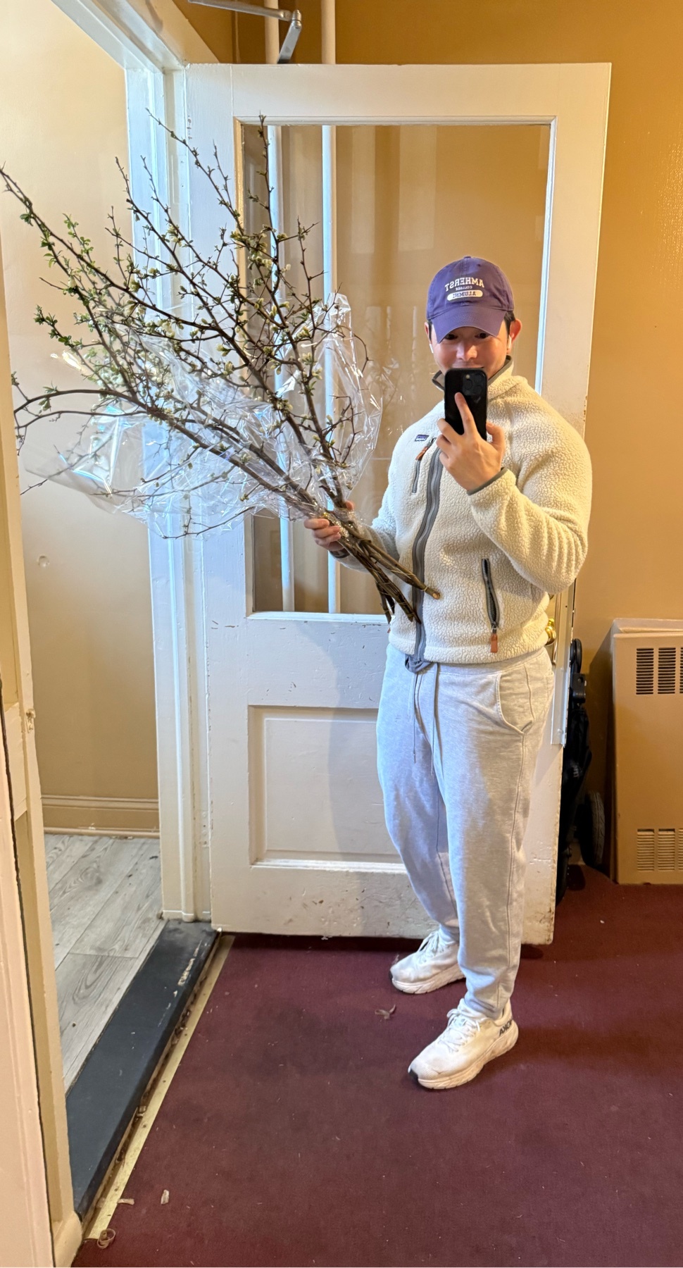 A man takes a selfie in a doorway while holding a large bouquet of branches with small white flowers. He is wearing a fleece jacket, sweatpants, and a baseball cap.