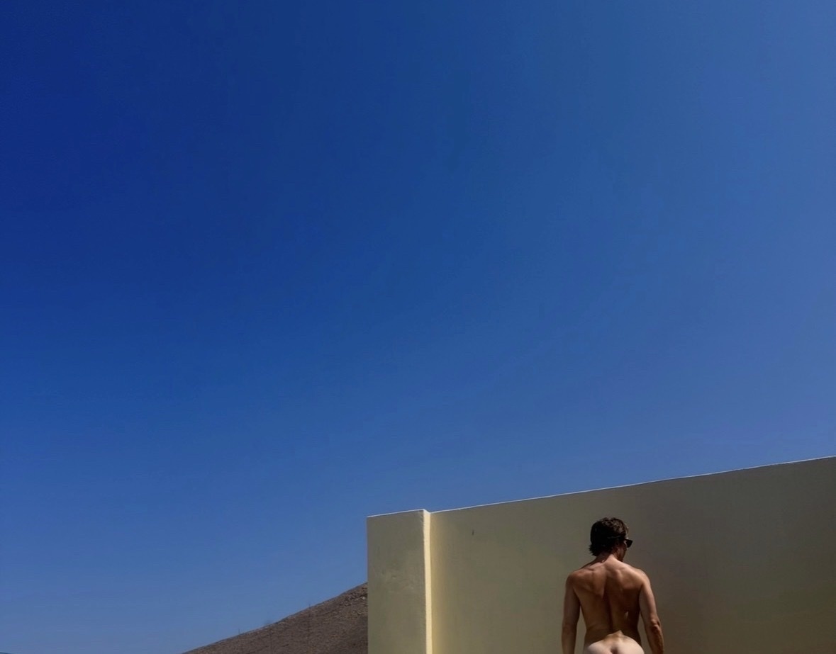 A shirtless man wearing sunglasses stands with his back to the camera, looking out at a clear blue sky and a light-colored wall.