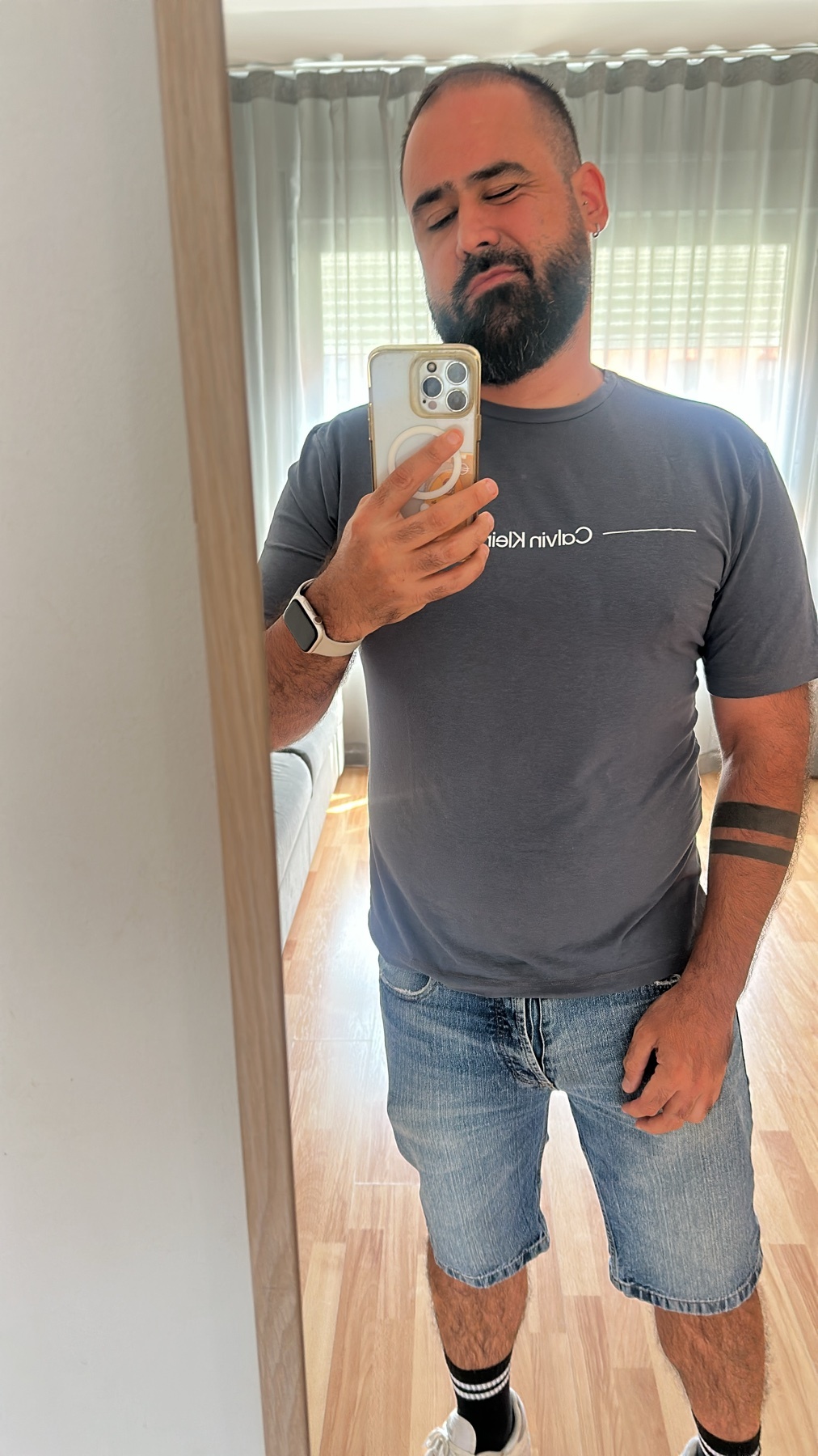 A man with a beard takes a selfie in a grey t-shirt and denim shorts. He is wearing white socks and sneakers.