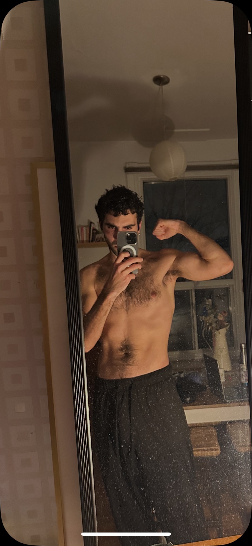 A man flexes his bicep in a mirror selfie. He is shirtless, showing off his muscular physique.