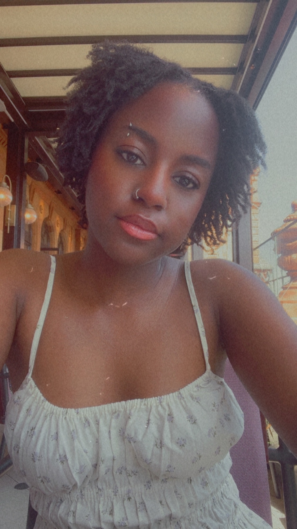 A young Black woman with locs and short hair poses for a selfie outdoors. She is wearing a light-colored floral print dress and has a nose ring and an earring.