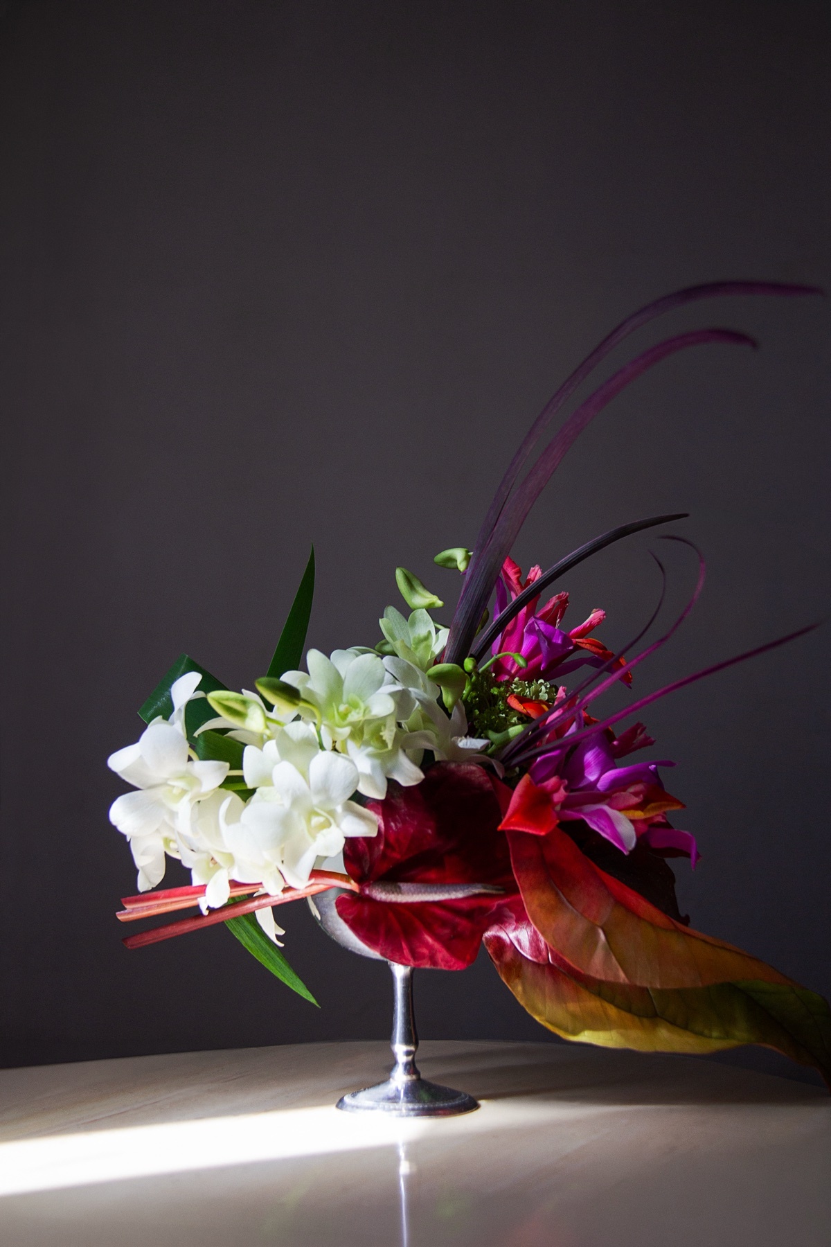A striking floral arrangement featuring white orchids and vibrant tropical flowers, artfully displayed in a silver goblet against a dark background.