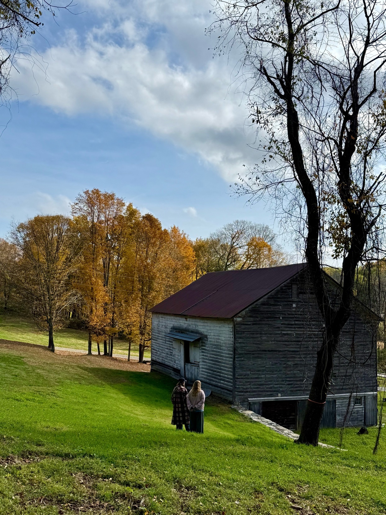A scenic autumn landscape featuring a wooden barn and colorful fall foliage. Two women are standing in front of the barn.