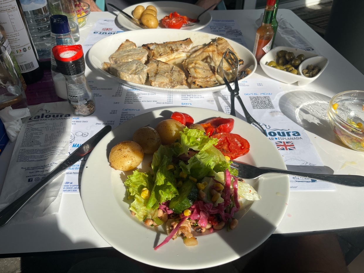 A close-up shot of a table laden with a delicious meal, featuring grilled fish, a fresh salad, boiled potatoes, and olives. The setting appears to be an outdoor restaurant, bathed in sunlight.
