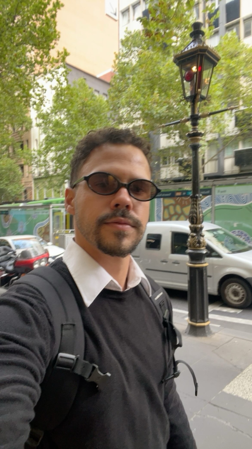 A man with sunglasses and stubble takes a selfie on a city street, with a lamppost and cars in the background.