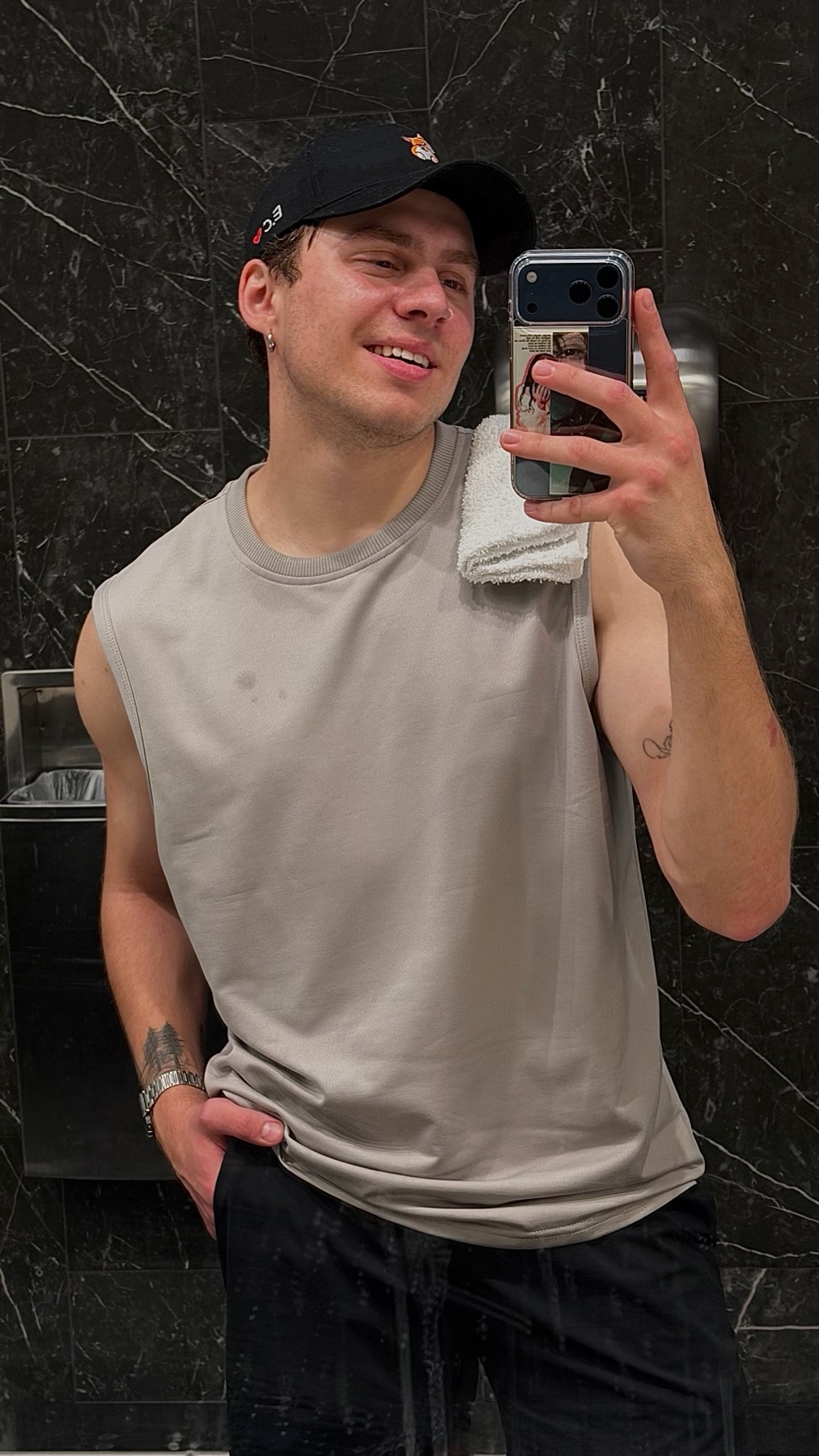 A man in a sleeveless shirt and baseball cap takes a selfie in a gym bathroom, smiling with a towel draped over his shoulder.