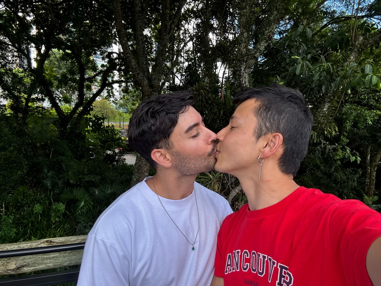 A selfie of two men kissing. One man is wearing a white shirt, and the other is wearing a red shirt.