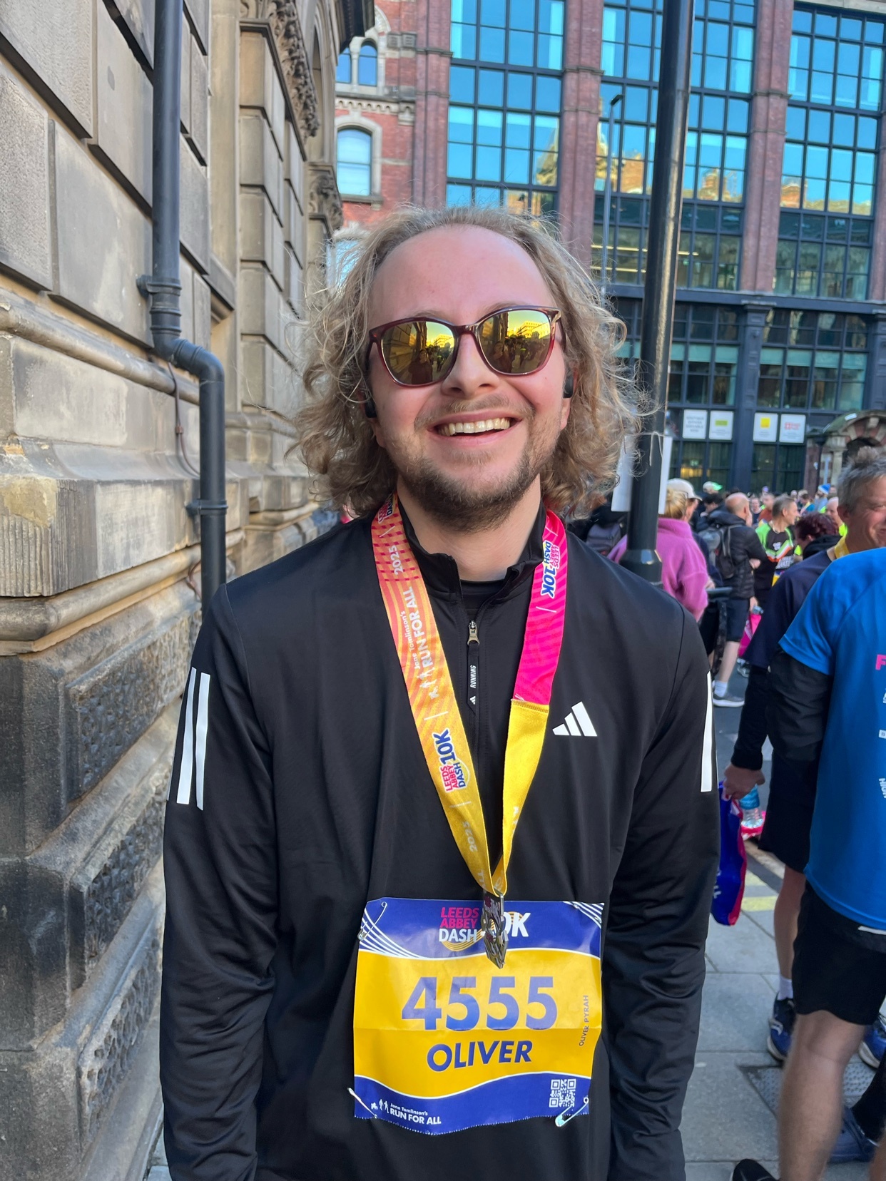 A man smiles while wearing a medal from a race. He is wearing sunglasses and a black Adidas jacket.