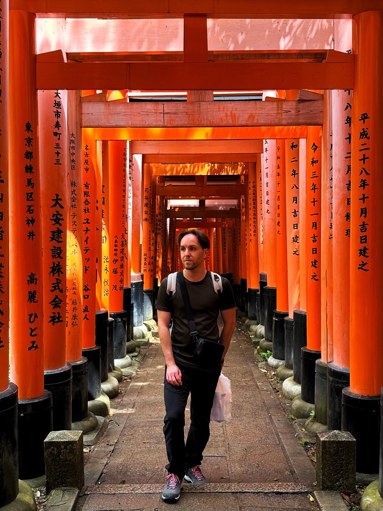 A man stands in the middle of a pathway lined with vibrant orange torii gates in Kyoto, Japan. The gates, inscribed with Japanese characters, create a tunnel-like effect, leading the eye into the distance.