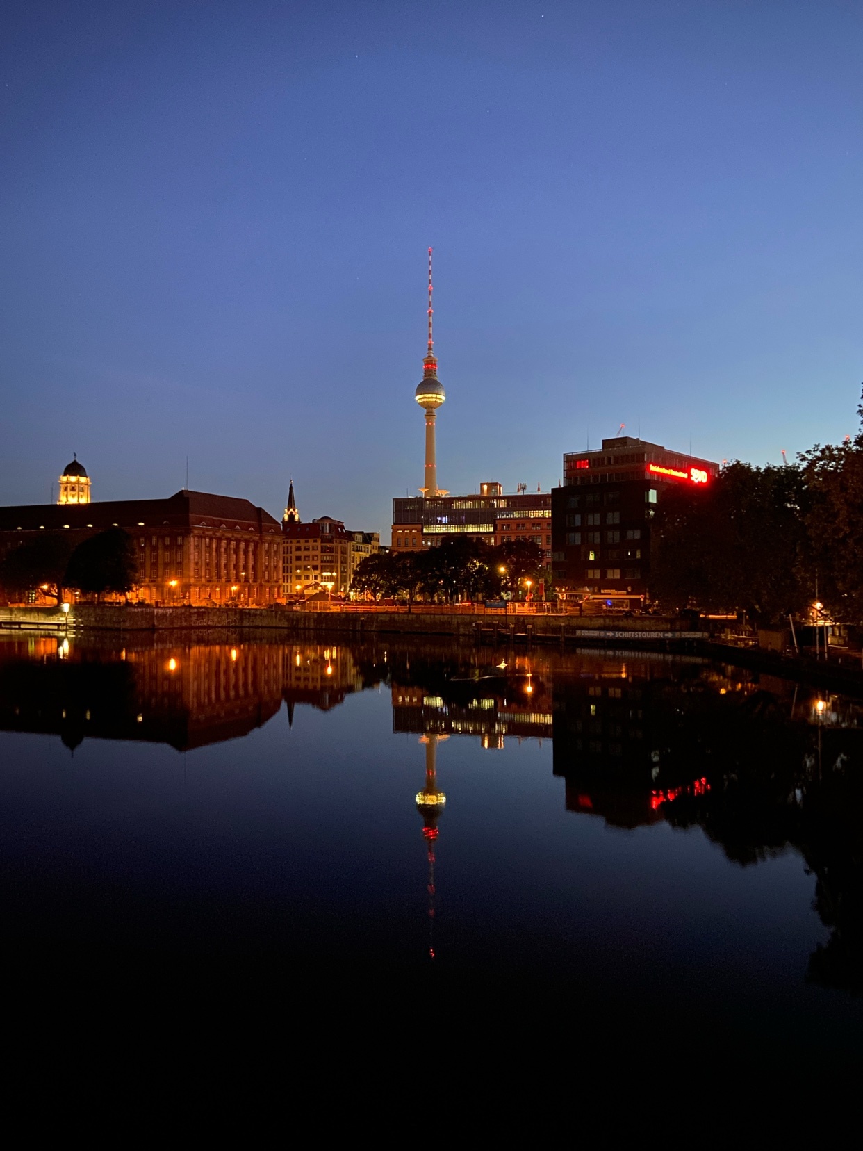 A nighttime cityscape is reflected in dark water. The iconic TV Tower stands tall in the center, illuminated against the deep blue twilight sky. Buildings on the left and right are also lit up, casting warm glows on the water's surface.