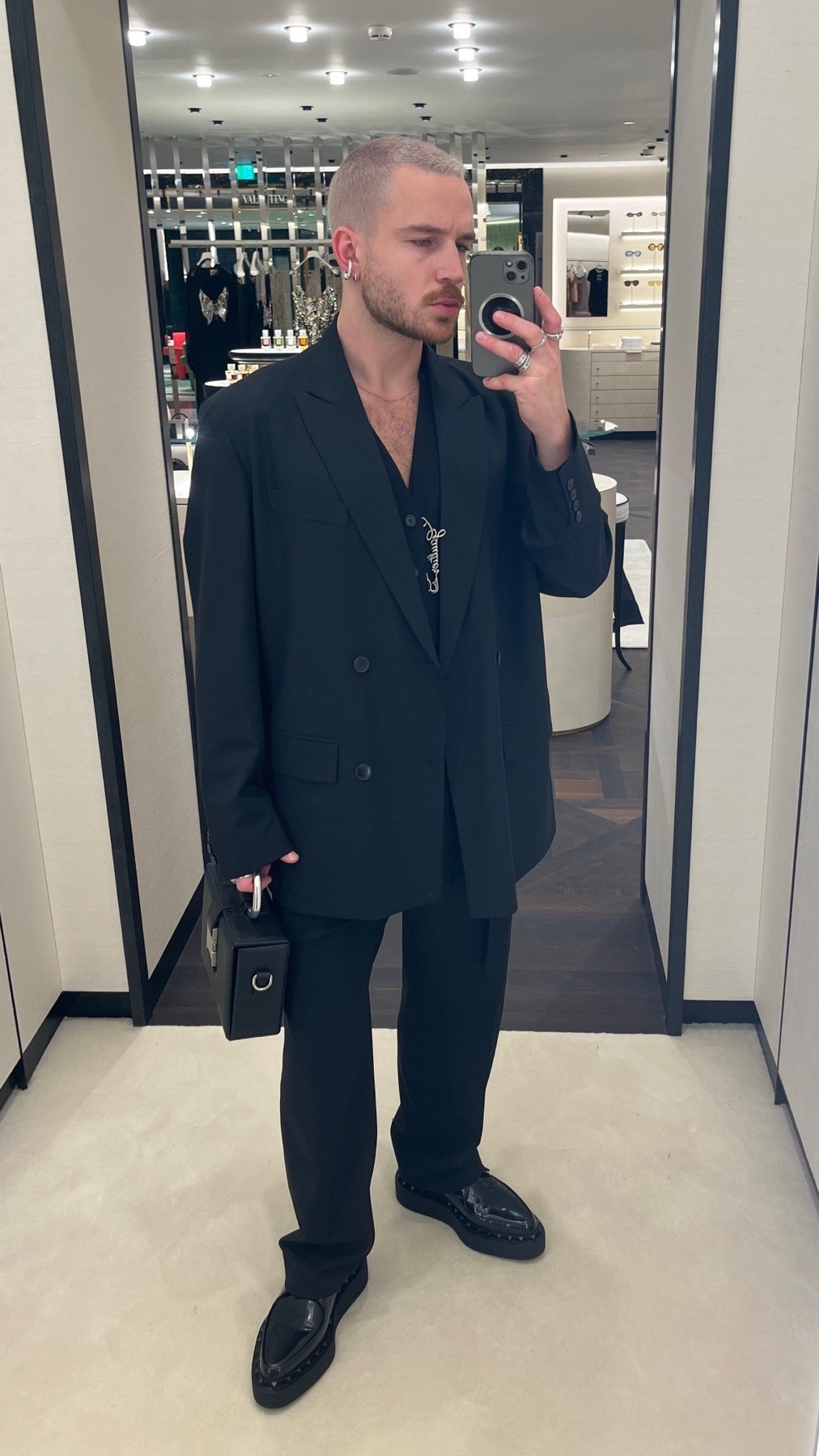 A man takes a mirror selfie in a black suit. The photo showcases a modern and stylish outfit.