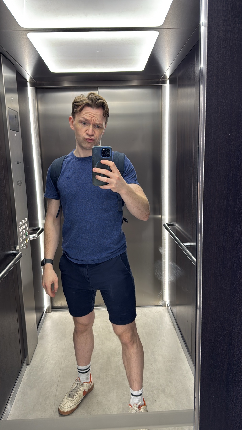 A man takes a selfie in a modern elevator, wearing a blue t-shirt, dark shorts, and a backpack.
